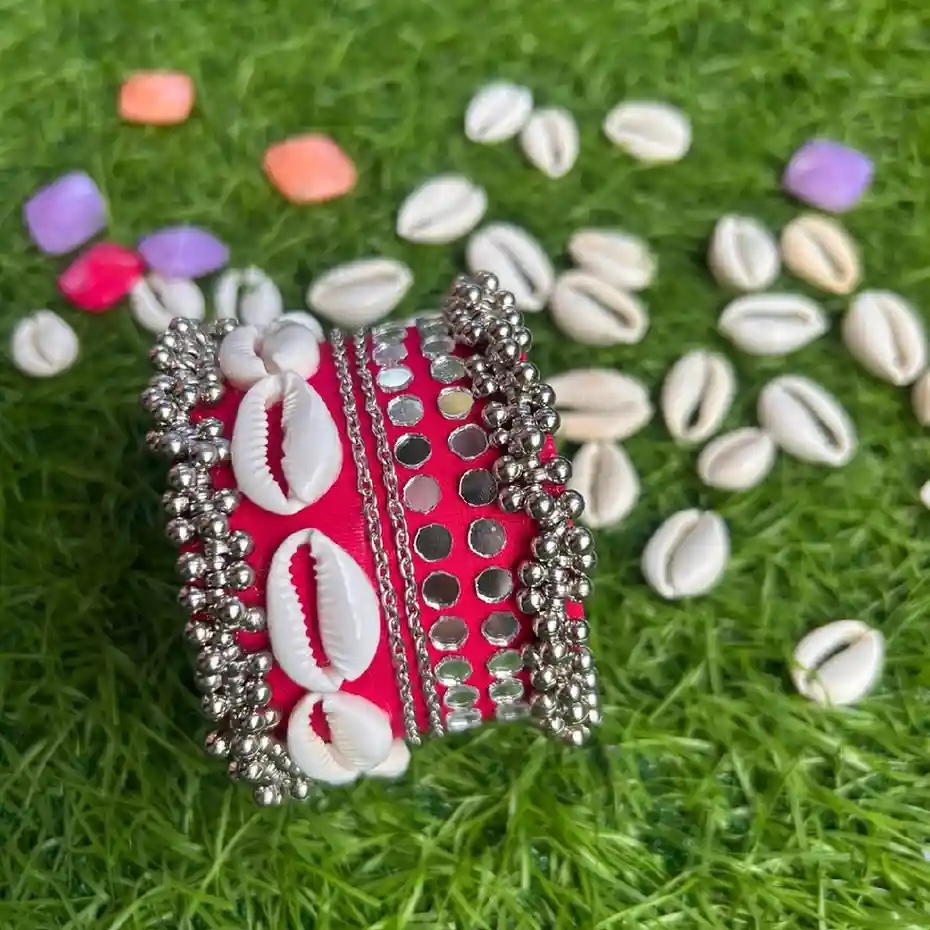 A handcrafted wide pink fabric handcuff with white cowrie shells, mirrors, and silver ghungroos displayed on a green grass background.