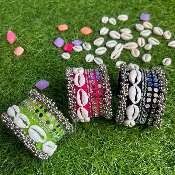 Three handcrafted fabric handcuffs in neon green, vibrant pink, and classic black, adorned with white cowrie shells, mirror work, and silver ghungroos, displayed on green grass.