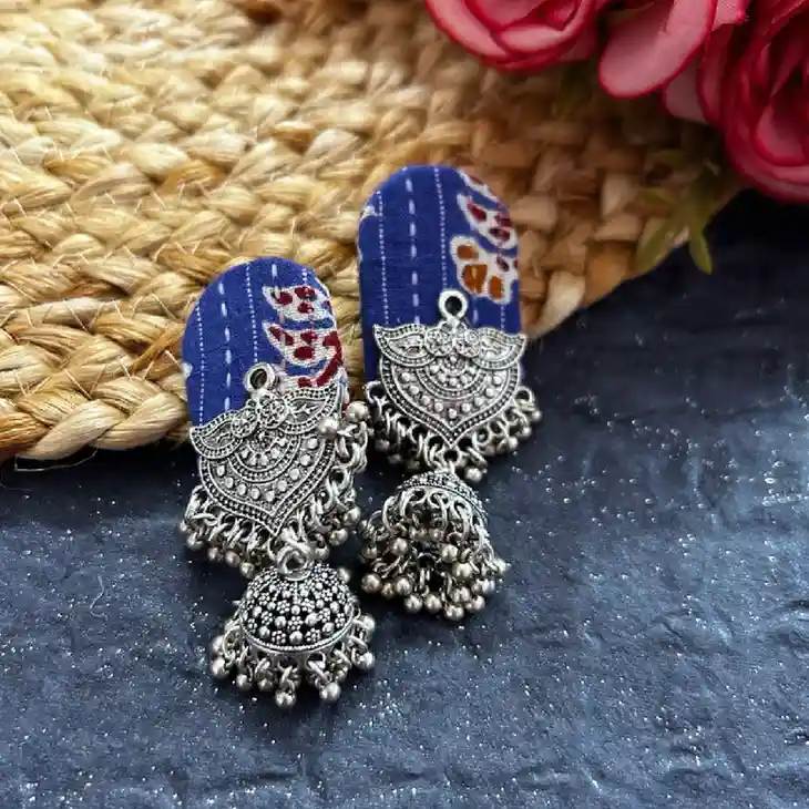 Close-up of indigo blue fabric-based oxidised silver jhumka earrings with intricate tribal design and dangling beads placed on a textured surface with pink flowers in the background