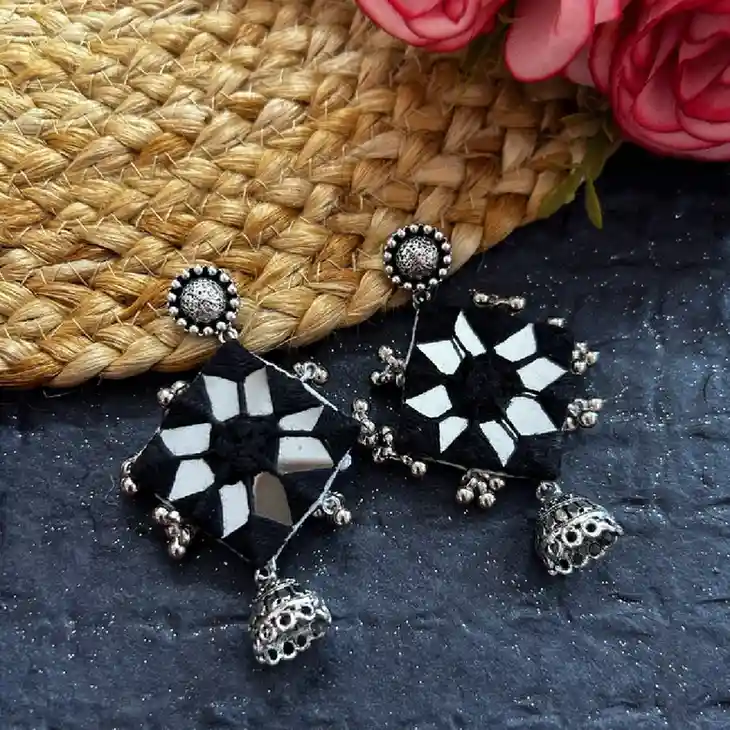 A pair of black fabric rhombus earrings featuring a central geometric star-mirror pattern, silver bead borders, and small oxidised silver jhumka drops on a dark textured background.