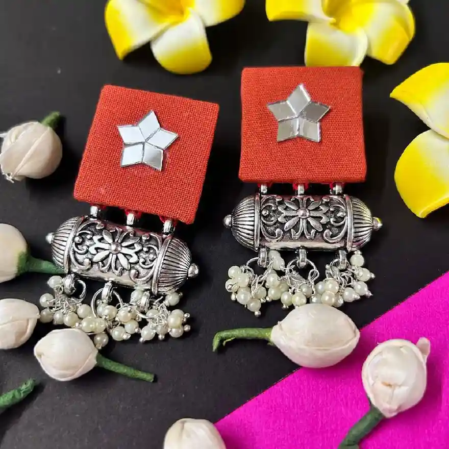 Pair of handcrafted orange fabric square earrings with star-shaped mirror work and silver oxidized floral beads with pearl clusters.