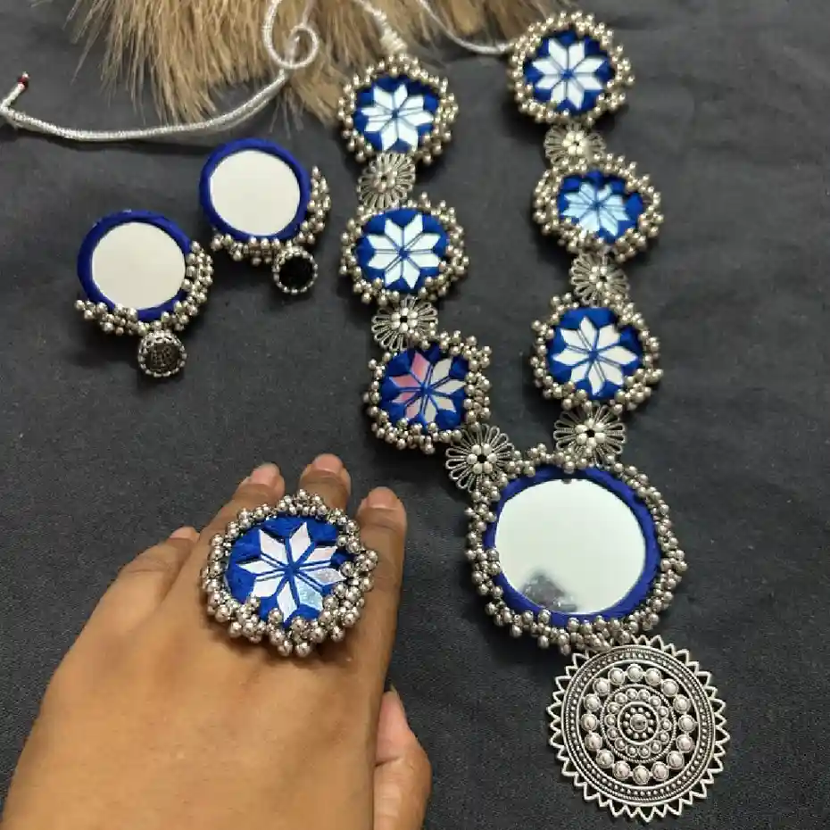 Handcrafted cobalt blue silk thread necklace with star-pattern mirror work, oxidized silver pendant, matching earrings, and statement ring on a grey background.
