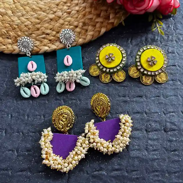 A collection of three pairs of artisanal fabric earrings: teal rectangular studs with pink and white cowrie shells, yellow circular mirror-work earrings with golden coins, and purple half-moon earrings heavily adorned with white seed pearls.