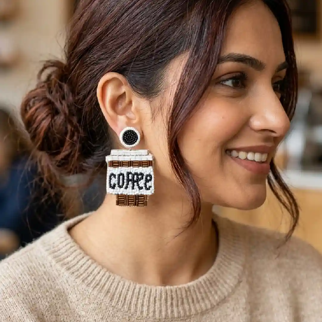 A model wearing handcrafted white and brown beaded statement earrings shaped like coffee cups with the word COFFEE in black beads, styled in a cozy cafe setting.