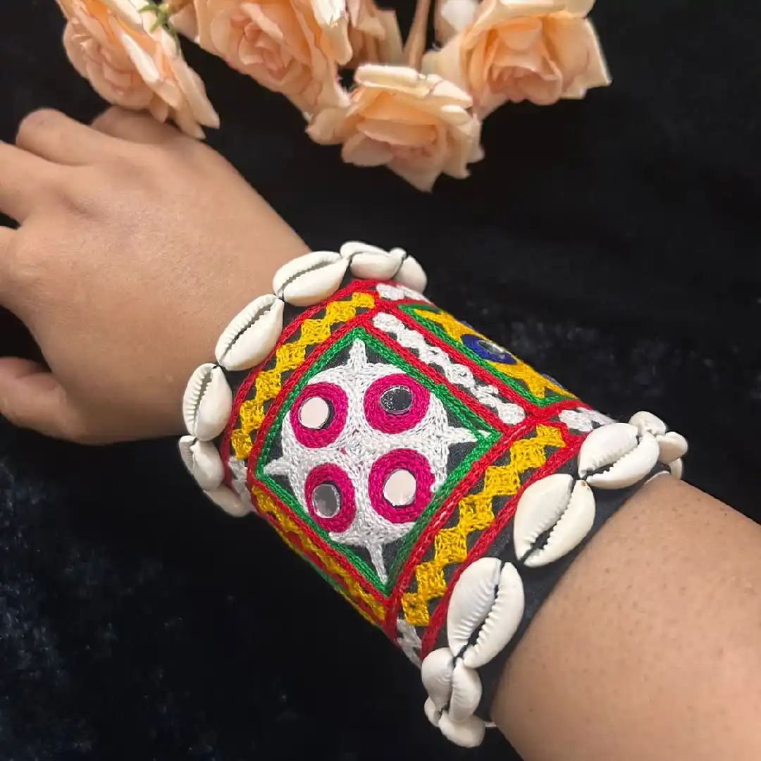 A person wearing a wide, handcrafted fabric cuff by Itsy Bitsy Kolkata featuring vibrant red, yellow, and green embroidery, real mirror work, and a border of white cowrie shells