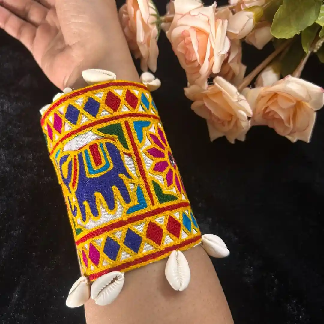 A hand wearing a wide yellow fabric cuff by Itsy Bitsy Kolkata, featuring a blue embroidered elephant, multi-color geometric patterns, and white cowrie shell drops.