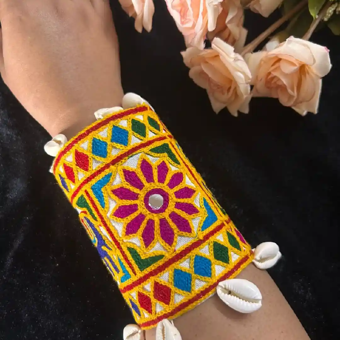 A handcrafted wide yellow fabric cuff by Itsy Bitsy Kolkata featuring a vibrant blue embroidered elephant, multi-color geometric Kutch patterns, and a border of white cowrie shells
