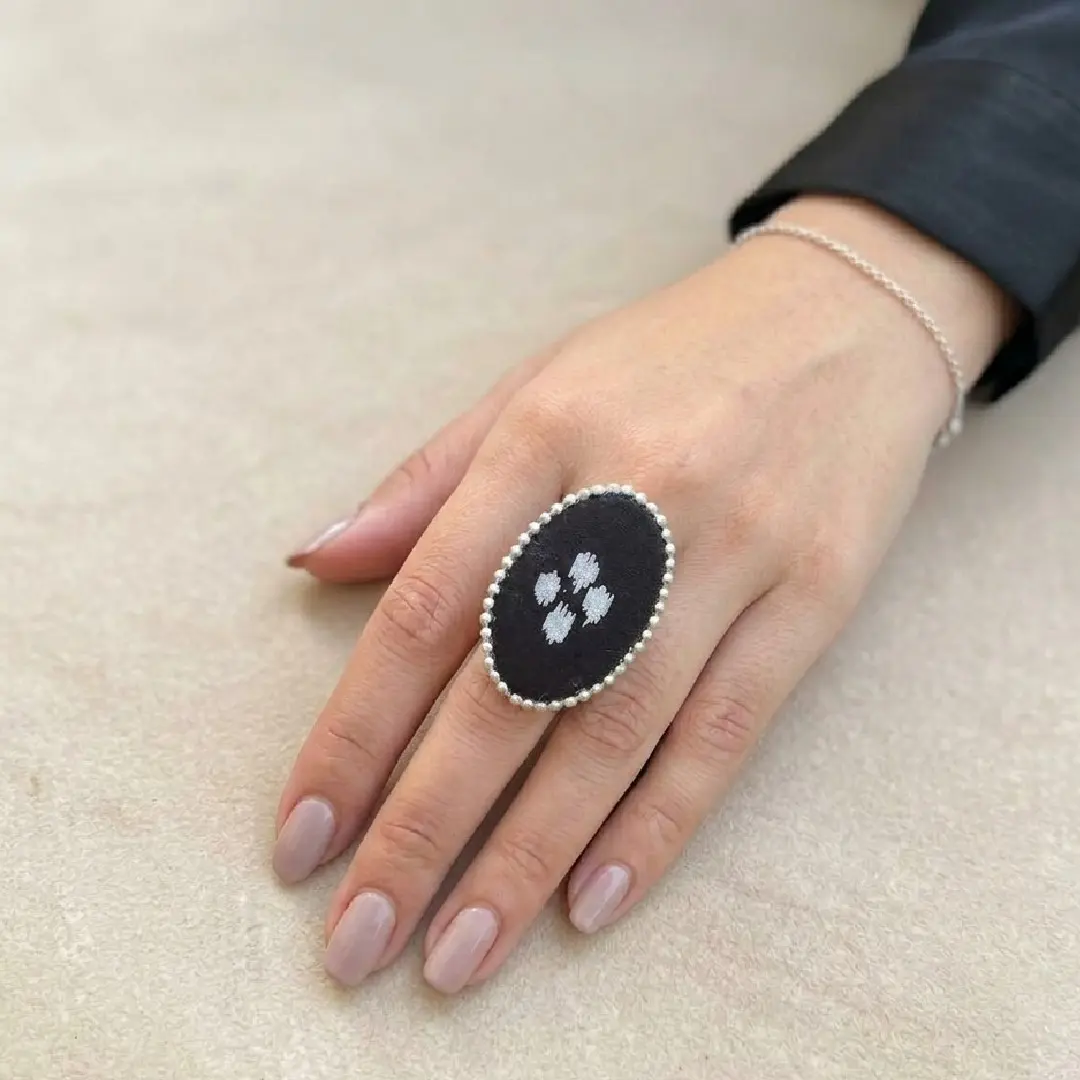 A model's hand featuring a single handcrafted black fabric oval statement ring with white embroidery and a silver bead border, styled against a clean, neutral minimalist background.