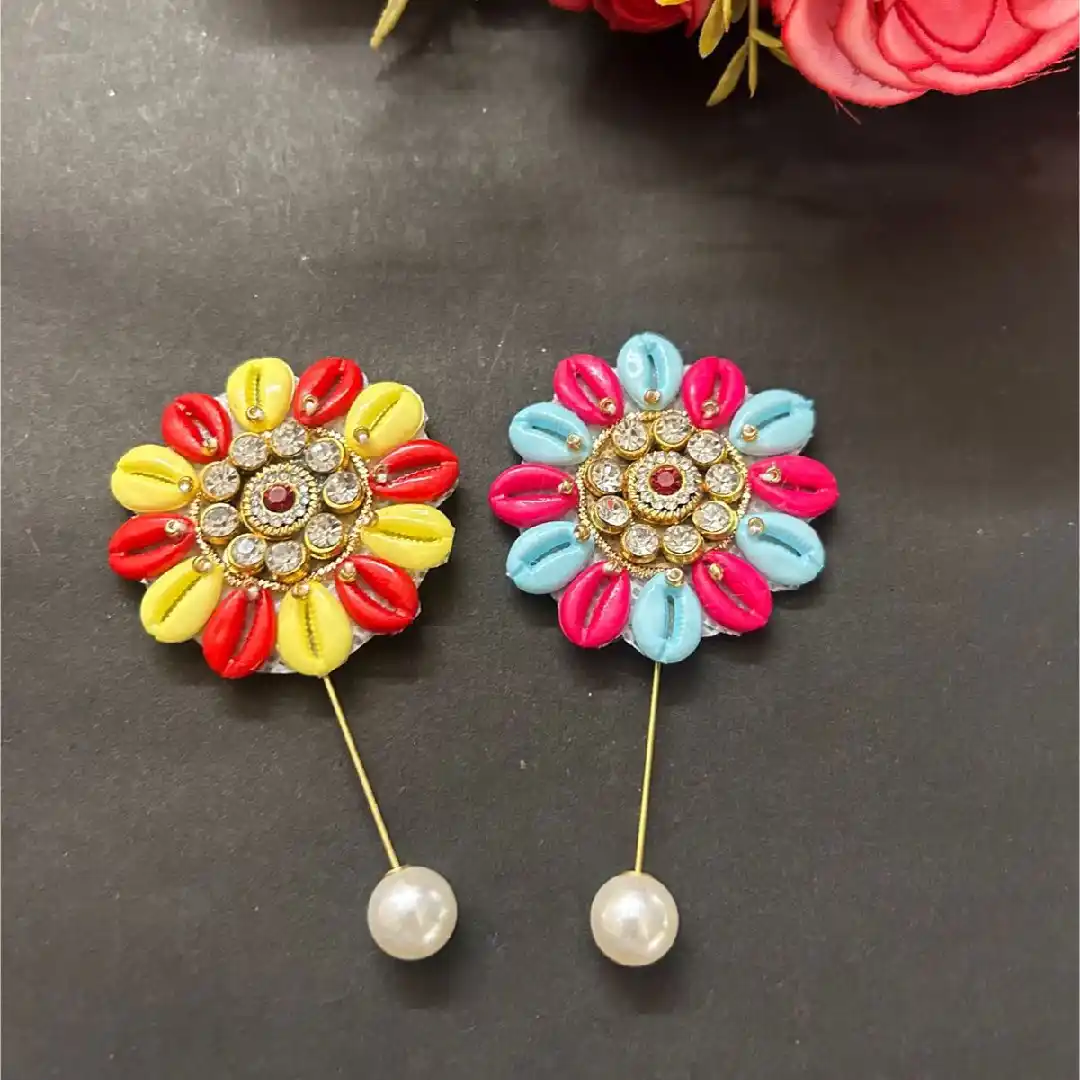 Two handcrafted floral pins made of red, yellow, blue, and pink cowrie shells with kundan centers and pearl drops.
