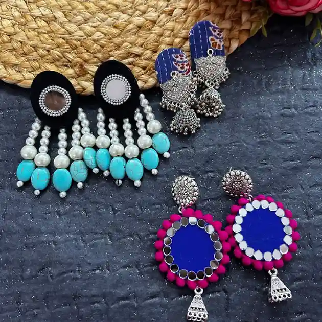A combo pack of three handcrafted earrings including turquoise pearl danglers, Ajrakh fabric jhumkas, and royal blue mirror-work drops on a textured grey background.