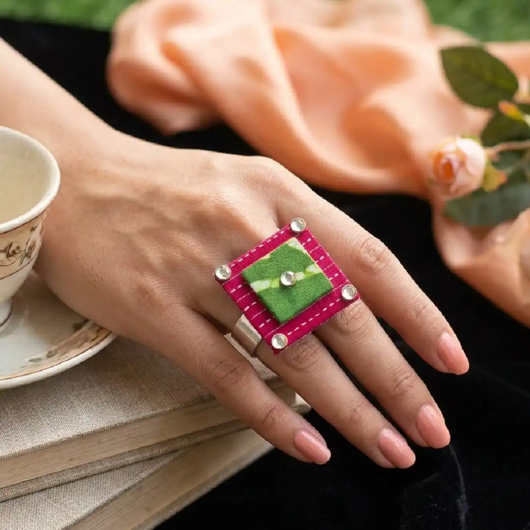 A model's hand with a soft purple manicure wearing a large handcrafted square statement ring made of pink and green fabric with crystal corner accents.