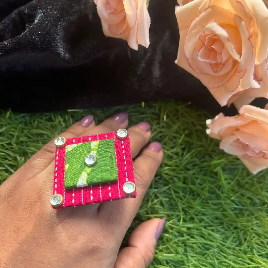 A person wearing a handcrafted oversized square statement ring made of pink and green fabric with white contrast stitching and sparkling stone corner accents.