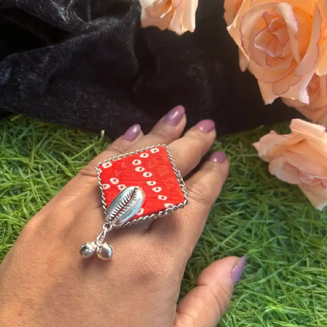 A person wearing a large handcrafted square statement ring made of red Bandhani fabric with a silver cowrie shell and hanging ghungroo beads.