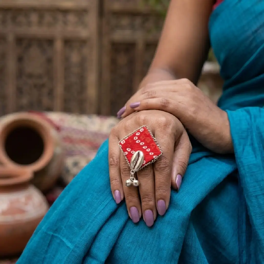 A professional lifestyle shot of a model styling an oversized handcrafted red Bandhani fabric statement ring with a silver cowrie shell and ghungroo beads, set against a soft-focus garden background.