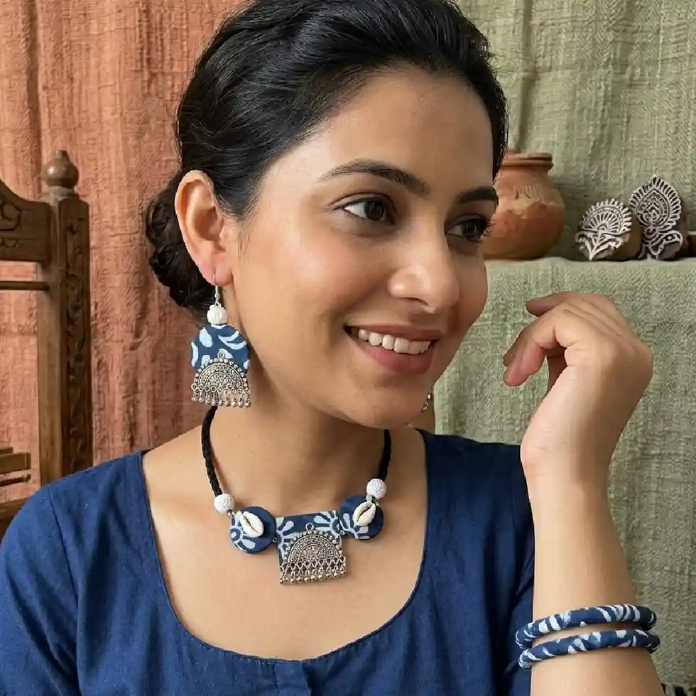 A model wearing an indigo blue Ajrakh fabric choker necklace with oxidized silver charms and cowrie shells, paired with matching earrings and fabric bangles against a soft, neutral indoor background.