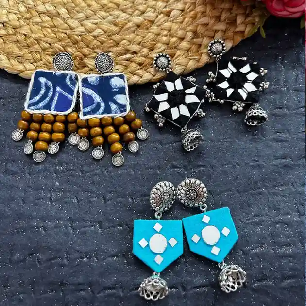 A collection of three pairs of handcrafted ethnic earrings featuring indigo fabric with wooden beads, black fabric with mirror stars, and turquoise fabric with jhumka drops.