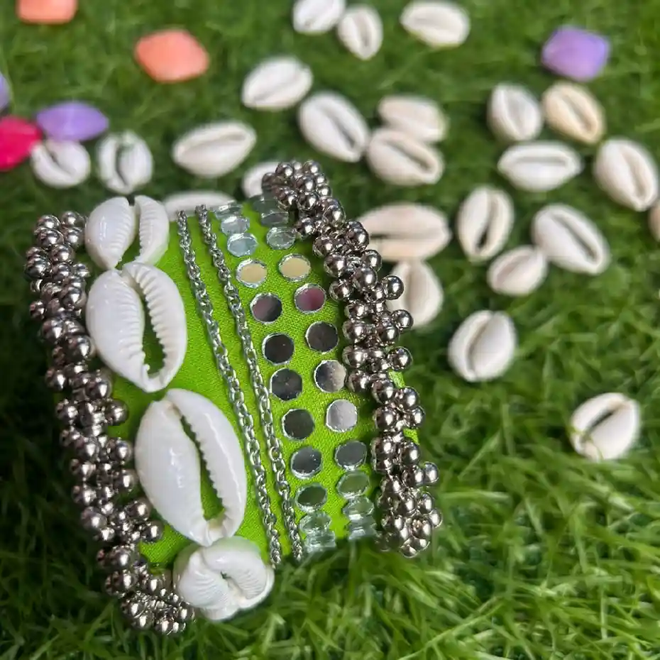 A handcrafted wide lime green fabric handcuff adorned with white cowrie shells, mirrors, and silver ghungroos displayed on a green grass background.