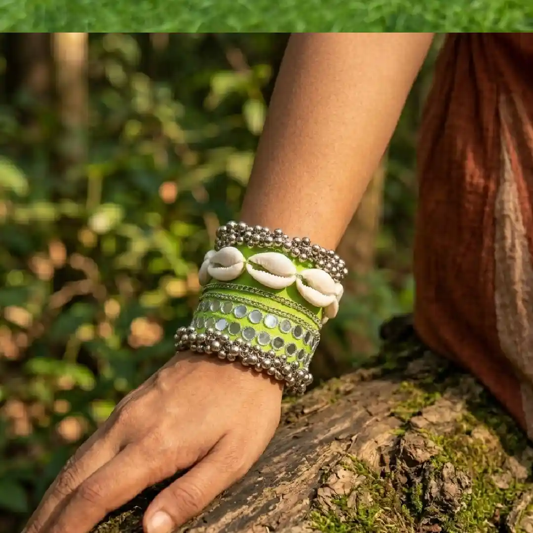 A close-up of a model's wrist wearing only a wide lime green handcrafted fabric handcuff with cowrie shells and mirror work against a blurred forest background.