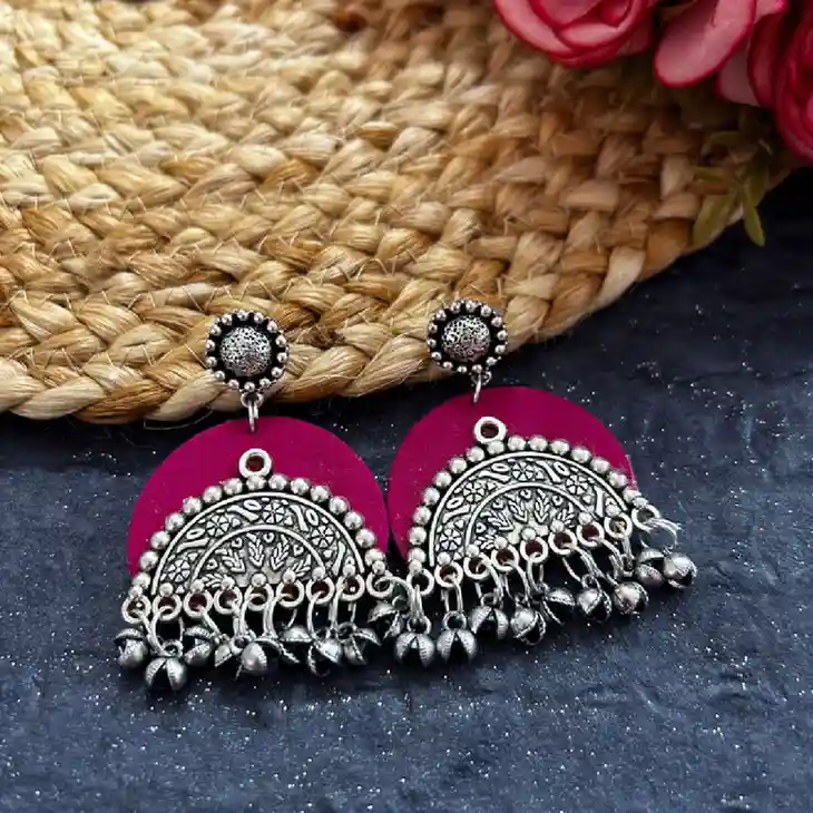 A pair of vibrant magenta fabric earrings featuring an intricate oxidised silver half-moon pendant and dangling metallic ghungroos on a dark textured background.
