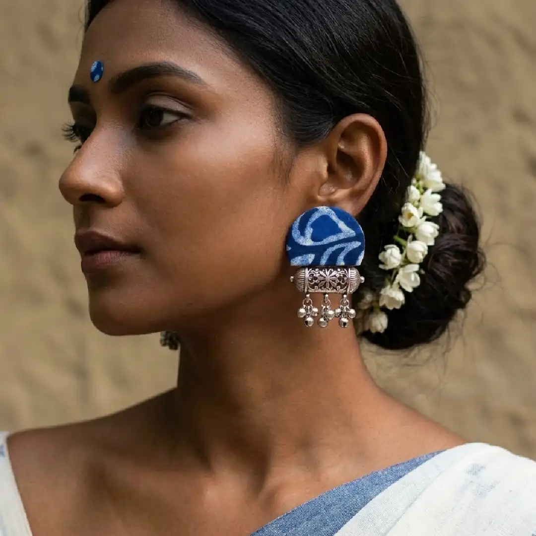 A model showcasing handcrafted Indigo Batik fabric stud earrings with oxidized silver dholak pendants and jingling ghungroo bells, styled with a traditional white cotton saree with a blue border.