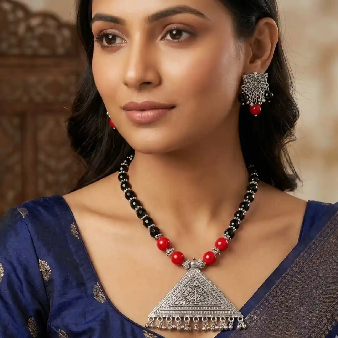 A model wearing a handcrafted oxidized silver tribal jewelry set with a large triangular pendant, matching earrings, and red and black beads, styled with a traditional black and maroon silk saree.