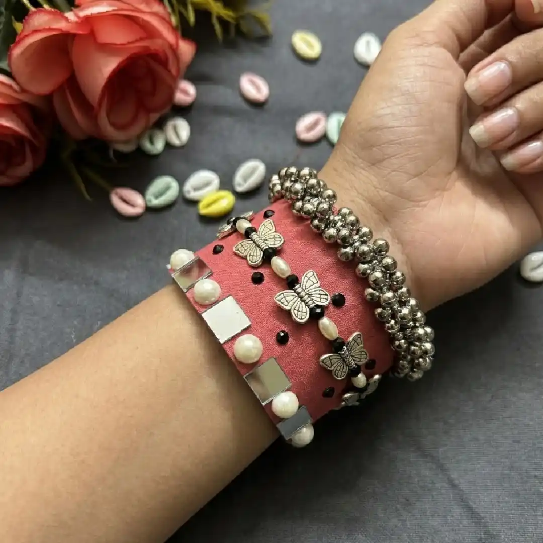 Handcrafted peach fabric handcuff bracelet from Itsy Bitzy Kolkata featuring silver butterfly charms, pearls, and mirror work, styled on a model's wrist with silver beads.