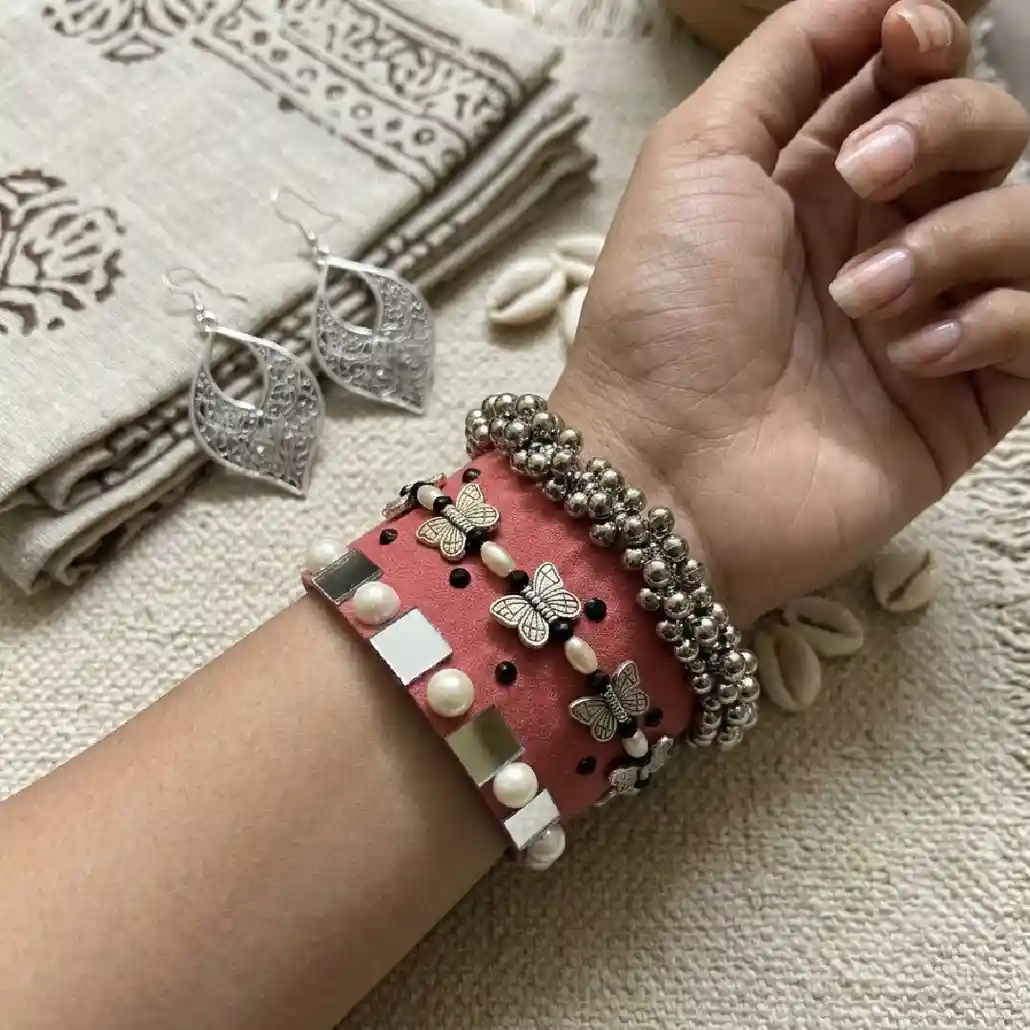 Model wearing a handcrafted peach fabric handcuff with silver butterfly charms, pearls, and mirror work from Itsy Bitzy Kolkata, styled with a silver beaded bracelet for a traditional ethnic look.