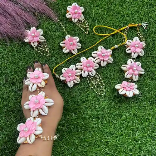 Complete pink floral and white cowrie shell bridal jewellery set featuring a necklace, earrings, maang tikka, and haathphool on a green grass background.