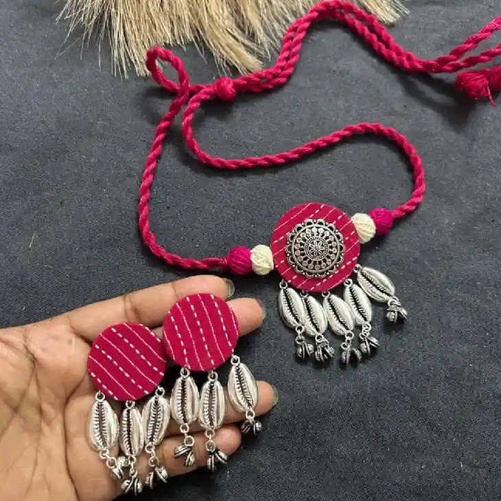 Handheld close-up of a rani pink fabric jewelry set from Itsy Bitzy Kolkata, showcasing matching circular earrings and a pendant with oxidised silver cowrie shells and ghungroo charms.