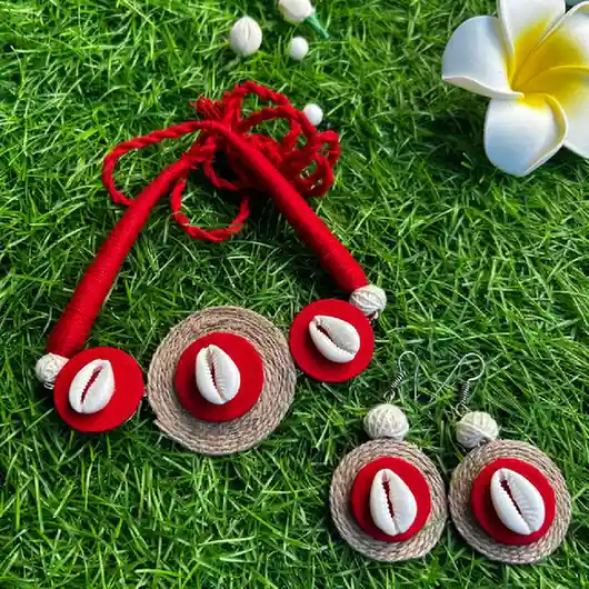 Handcrafted ethnic jewelry set featuring a red and beige jute choker necklace and matching drop earrings adorned with white cowrie shells, displayed on a green grass background.
