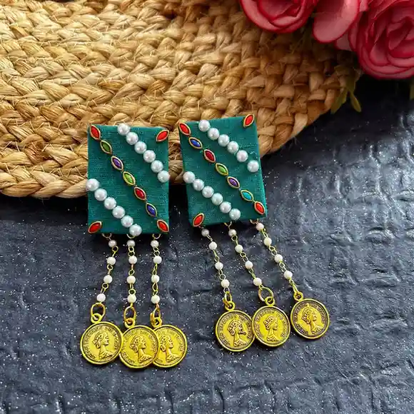 A pair of artisanal teal fabric earrings featuring rectangular bases adorned with diagonal pearl bead patterns, finished with three cascading chains of small white pearls and engraved golden coin charms on a dark textured background