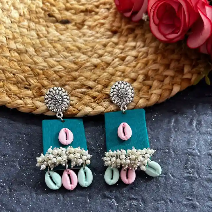 A pair of rectangular teal fabric earrings featuring central pink cowrie shells, white pearl bunch clusters, and hanging pink and mint cowrie shells on oxidised silver floral studs.