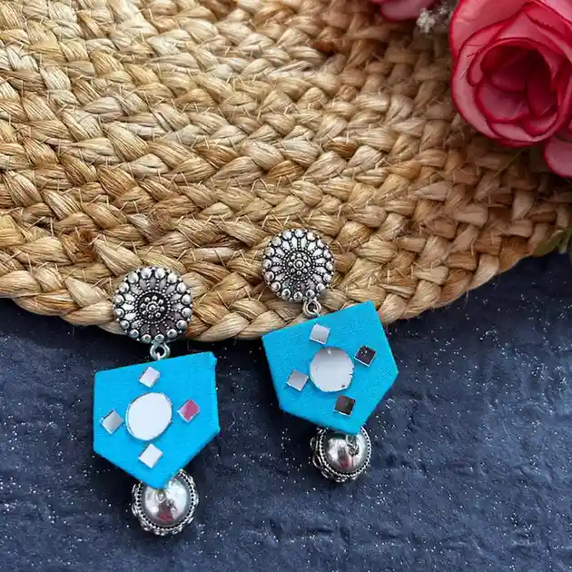 A pair of turquoise blue pentagonal fabric earrings featuring central circular and diamond-shaped mirror work, attached to intricate oxidised silver floral studs with a silver bead drop.