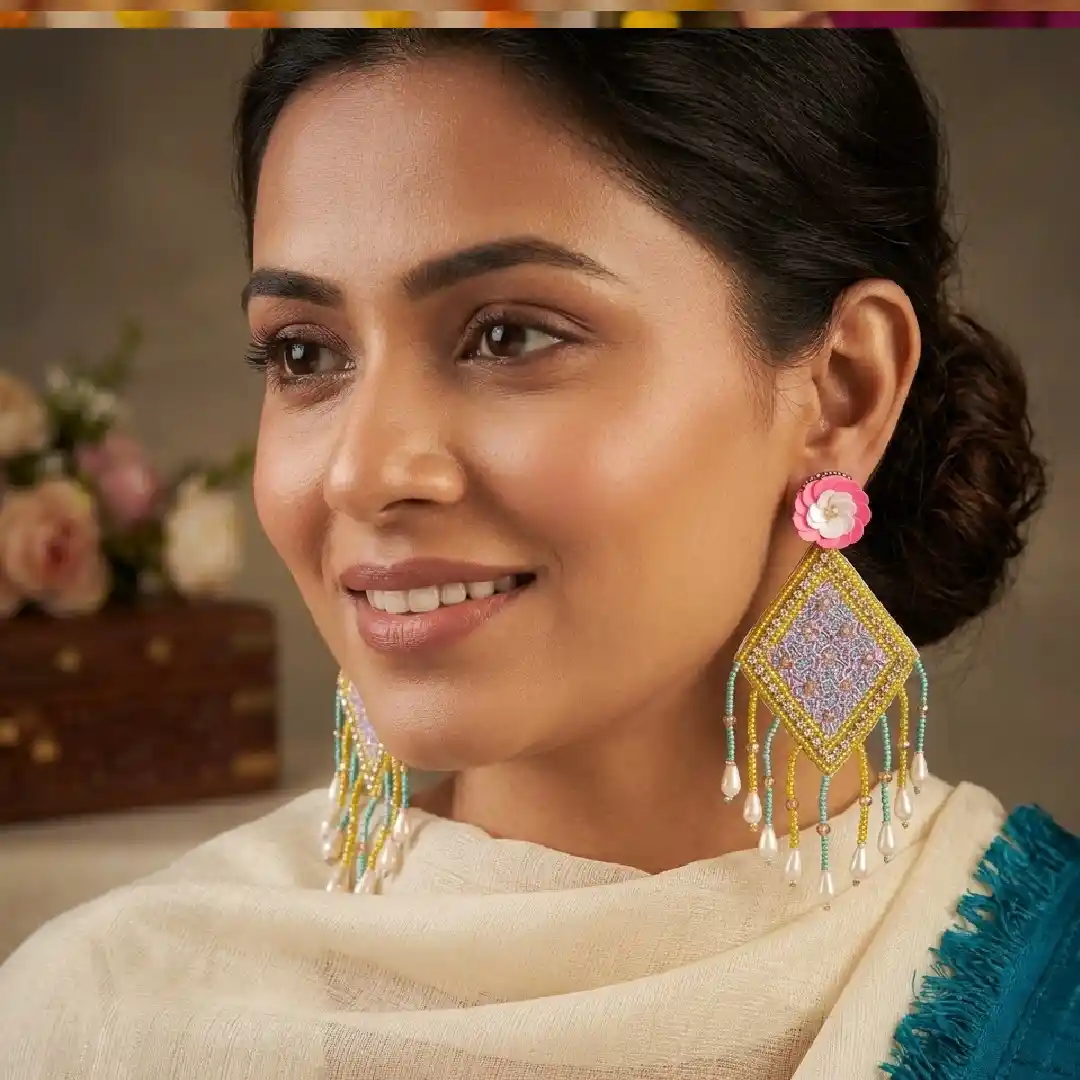A close-up portrait of a woman wearing large, handcrafted diamond-shaped beaded statement earrings featuring pink flower motifs, yellow and purple geometric patterns, and pearl fringe accents.