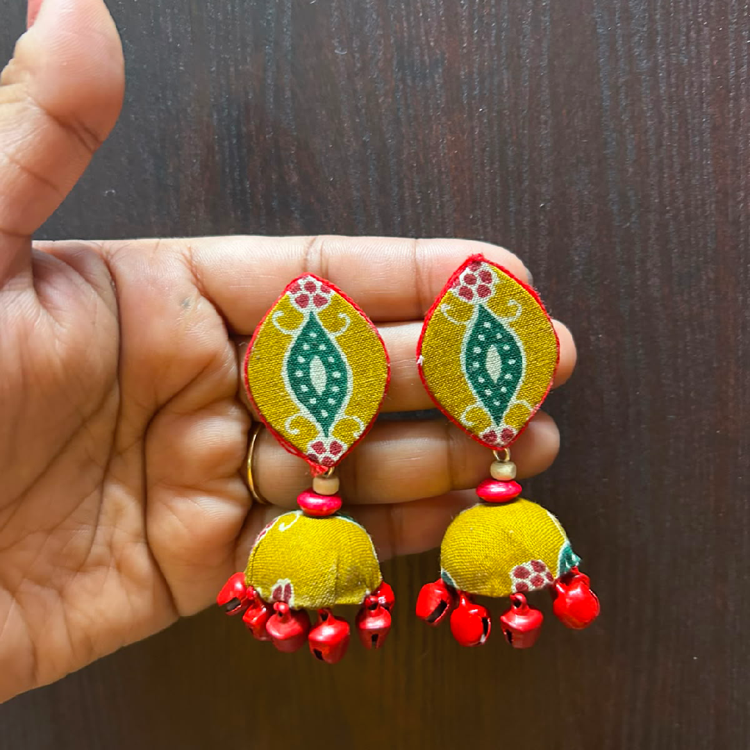 Handmade ethnic jhumka earrings featuring leaf-shaped yellow fabric studs with white and green patterns, a yellow dome drop, and hanging red metal ghungroo bells.